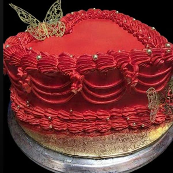 A heart-shaped red cake with intricate frosting, gold edible pearls, and a gold butterfly decoration.