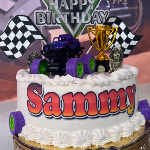 White birthday cake with "Sammy" in red and orange, topped with a monster truck and racing flags.