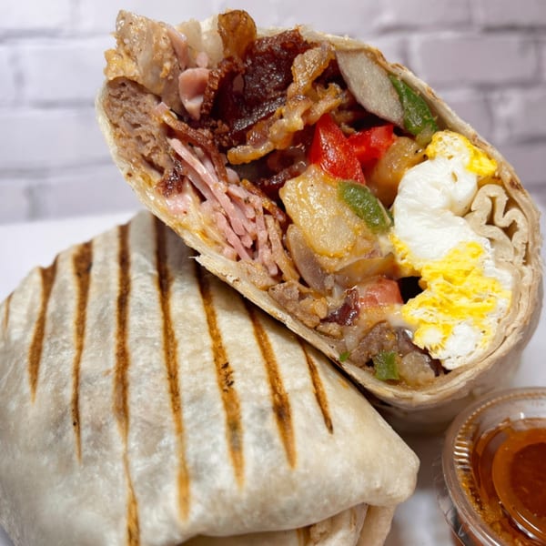 A grilled breakfast burrito, cut in half, revealing eggs, bacon, ham, and vegetables.