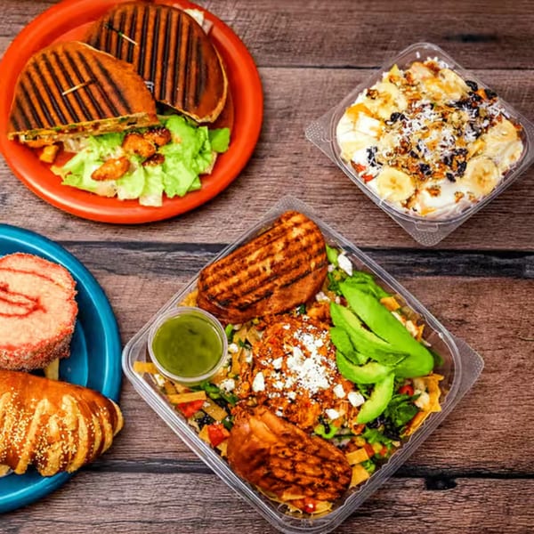 Assortment of food including a panini, salad, fruit bowl, and pastry on a wooden table.