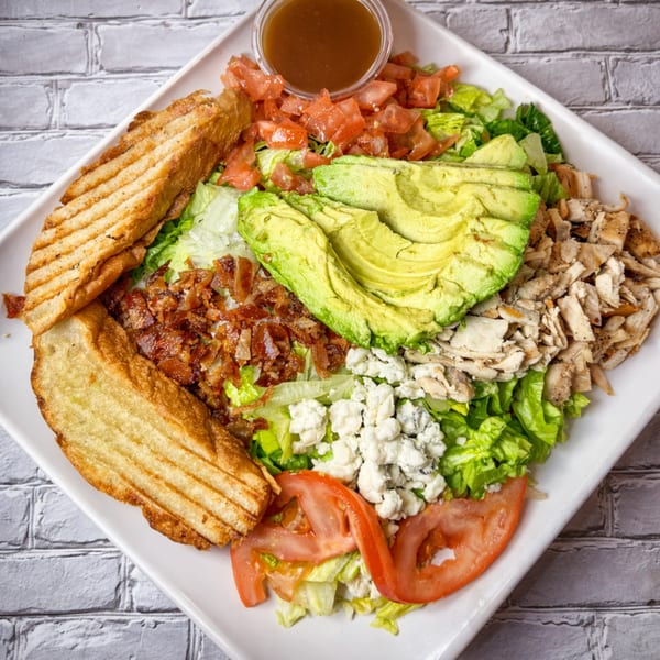 A vibrant salad with grilled bread, avocado, bacon, chicken, blue cheese, and tomatoes.